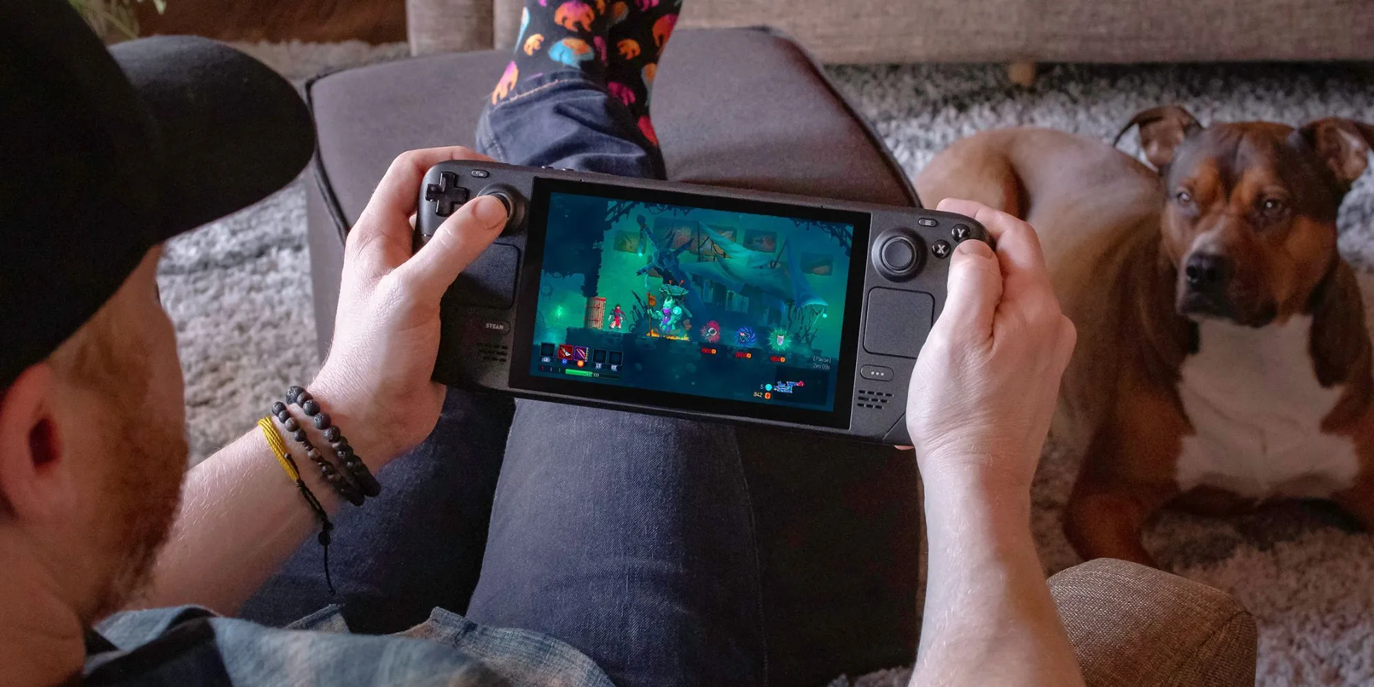Người đàn ông thư giãn trên sofa chơi game Steam Deck cùng chú chó cưng bên cạnh
